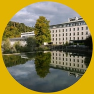 Gebäude am Flussufer mit Bäumen und Spiegelung im Wasser.