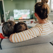 Person auf Sofa mit zwei Kindern, schaut TV.
