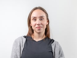 Person mit langen Haaren, grauem Pullover und schwarzen T-Shirt vor hellem Hintergrund.