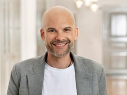 Mann mit Glatze in grauem Blazer lächelt.