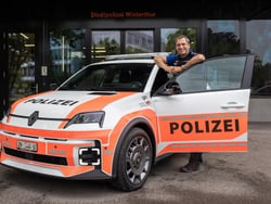 Person neben einem Polizeiauto mit geöffneter Tür vor einem Gebäude.
