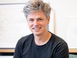 Person mit grauem Haar in schwarzem T-Shirt lächelnd.