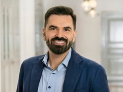 Mann mit Bart in blauem Sakko lächelt in Innenraum.