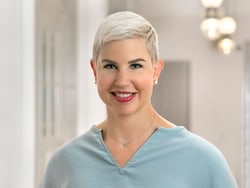 Person mit kurzen, blonden Haaren und blauem Oberteil lächelt.