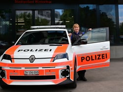 Person in Uniform steht neben einem Polizeiauto vor einem Gebäude.