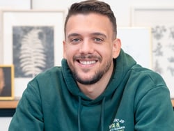 Ein lächelnder Mann in einem grünen Hoodie sitzt vor einer Wand mit gerahmten Bildern.