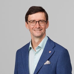 Stadtrat Andreas Geering lächelnd in blauem Anzug mit Brille vor grauem Hintergrund.