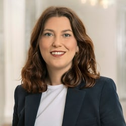 Person mit braunen Haaren und blauem Blazer lächelt.