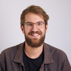 Mann mit Brille und Bart lächelt vor grauem Hintergrund.