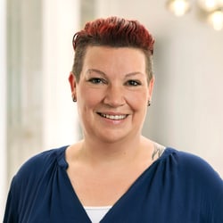 Person mit kurzem dunklem Haar und blauem Oberteil lächelt in die Kamera.