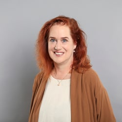 Frau mit rotem Haar und braunem Pullover vor grauem Hintergrund.
