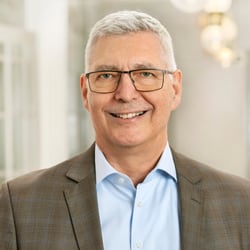 Mann mit Brille und Jackett lächelt in einem hellen Raum.