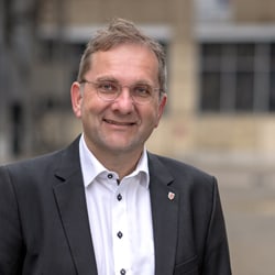 Mann in Anzug mit Brille lächelt draussen.