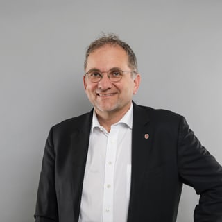 Person mit Brille in Anzug vor grauem Hintergrund.