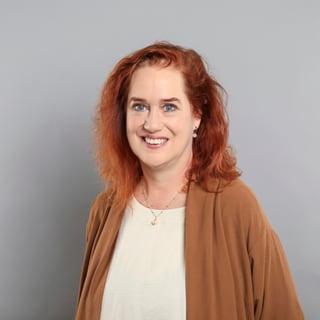 Frau mit rotem Haar und braunem Pullover vor grauem Hintergrund.