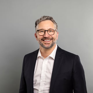 Mann mit Brille im Anzug vor grauem Hintergrund.