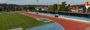 Laufbahn und Fussballfeld im Freien mit Tribünen und Gebäuden im Hintergrund.
