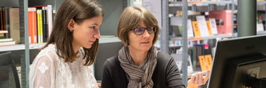 Zwei Personen sitzen vor einem Computer in einer Bibliothek.