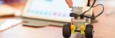 Person zeigt auf einen Lego Roboter auf einem Tisch vor Tablet.