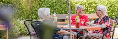Drei ältere Frauen sitzen lachend an einem Tisch in einem Restaurant. Sie trinken gekühlte Getränke unter einem Sonnnschirm.