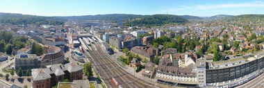 Panoramablick auf eine Stadt mit Bahngleisen und Hügeln im Hintergrund.