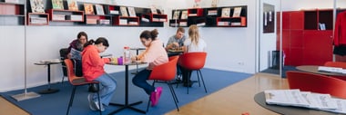 Menschen sitzen an Tischen in einem modernen Leseraum mit Regalen.