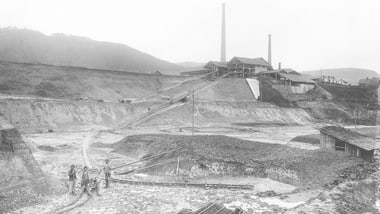 Historische Industrieanlage mit Schornsteinen und Hügeln, vier Personen im Vordergrund.