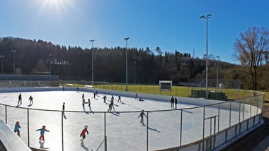 Eislaufbahn im Freien bei Sonnenschein, umgeben von Bäumen.