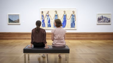 Zwei Personen sitzen auf einer Bank in einer Kunstgalerie, umgeben von Gemälden.