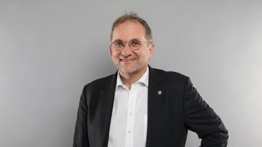 Person mit Brille in Anzug vor grauem Hintergrund.
