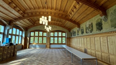 Raum mit gewölbter Holzdecke und grossen Fenstern, langer Tisch an der Wand.