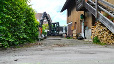 Bagger in einem Hof neben einem Holzhaus und Holzstapeln.