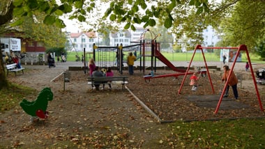 Spielplatz mit Kindern, Schaukeln und Rutschen, umgeben von Bäumen.