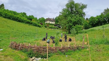 Menschen arbeiten in einem Garten auf einem Hügel mit Gebäude im Hintergrund.