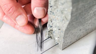 Hand hält Fasern neben einem Betonblock mit herausragenden dünnen Stäben.