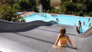 Kind auf Wasserrutsche im Freibad, mit Menschen im Pool und Landschaft im Hintergrund.