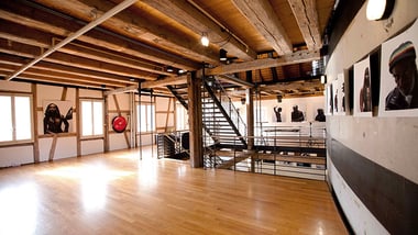 Lichtdurchfluteter Raum mit Holzboden und sichtbaren Balken, Treppe im Hintergrund.