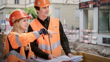 Zwei Bauarbeiter in orangefarbenen Westen und Helmen betrachten Baupläne.