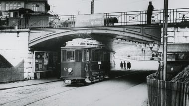 Schwarzweissfoto mit Strassenbahn unter einer Brücke und Menschen im Hintergrund.