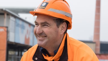 Person in orange Bauarbeiterjacke und Helm lächelnd auf Baustelle.