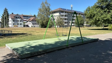 Leerer Spielplatz mit Schaukel und Gebäuden im Hintergrund.
