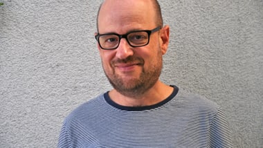 Mann mit Brille und gestreiftem Hemd vor grauer Wand.