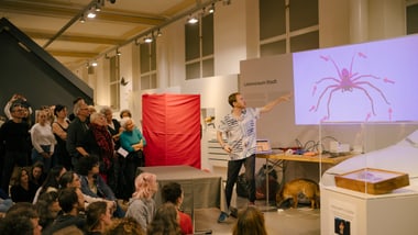Person hält Vortrag zu Spinnen vor Publikum in einem Ausstellungsraum.