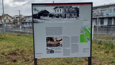 Informationsschild neben Bahngleisen auf grasbewachsenem Bereich.