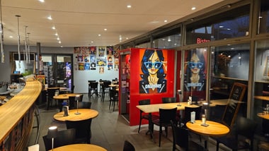 Innenansicht eines leeren Cafés mit Holzstühlen und Tischen, bunte Wandkunst.