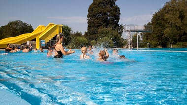 Menschen schwimmen im Pool mit gelber Rutsche im Hintergrund.