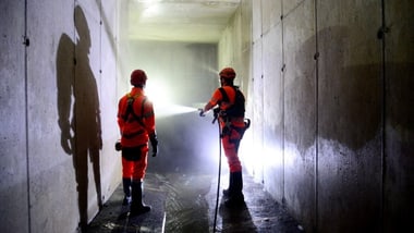 Zwei Personen in Schutzkleidung in einem dunklen Tunnel mit Taschenlampen.