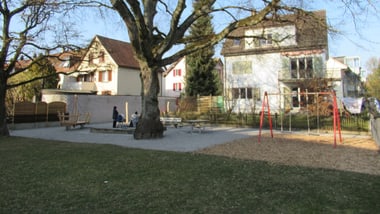 Spielplatz mit Bäumen und Wohnhäusern im Hintergrund.
