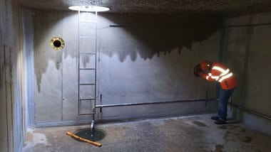 Person in leuchtender Arbeitskleidung in einem leeren Betonraum mit Leiter.
