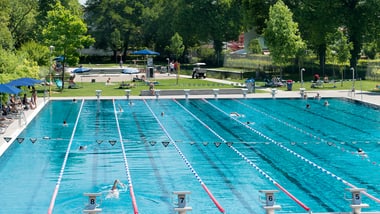 Freibad mit Schwimmern und Bäumen im Hintergrund.